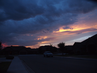 [View down a street with blue-grey storm clouds overhead mainly to the left. Beyond those in the distance just above the rooflines the sky is pink. To the right there is blue sky which then melds into the purple with splashes of yellow and pink as the sun catches the edges of those clouds.]